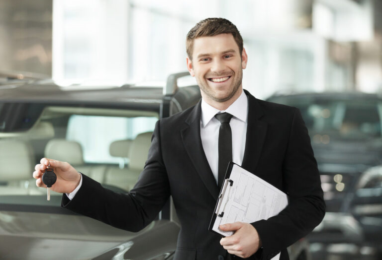 a male car dealer smiling while holding clipboard and key